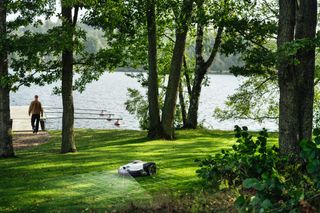 A robotic lawn mower operating on a grassy area near a lake, with a person walking in the background.