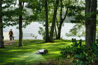 A robotic lawn mower operating on a grassy area near a lake, with a person walking in the background.