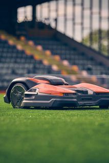 Роботизирана косачка за трева, работеща на спортно игрище с празни трибуни на заден план.