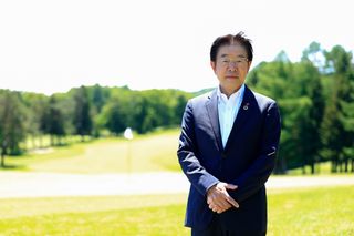 Person standing with hands clasped on a sunny golf course with trees in the background.