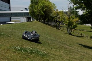 建物や木の近くの芝が茂った斜面で動作するハスクバーナのロボット芝刈機。