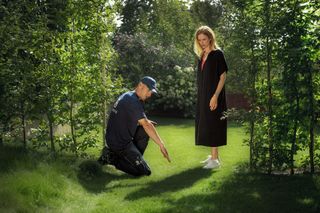 Una persona arrodillada en el césped de un jardín, interactuando con otra persona de pie.