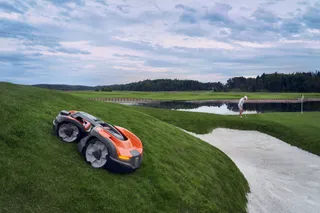 A robotic lawn mower operating on a golf course slope near a sand bunker and a golfer.