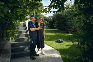 Två personer som står på en uteplats och observerar en robotgräsklippare som arbetar i en trädgård.