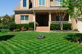 Robotic lawn mower operating on a striped lawn in front of a suburban house.