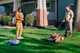 Husqvarna robotic lawn mower on grass while a person pushes a mower in a residential front yard.