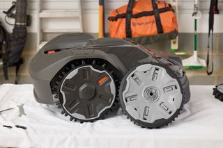 Side view of a robotic lawn mower on a table, with tools and a bag in the background.