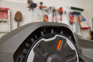 Close-up of a robotic lawn mower wheel with gardening tools hanging on a wall in the background.