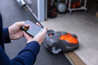 Une personne contrôlant une tondeuse à gazon robotisée à l’aide d’une application pour téléphone intelligent près d’un garage.