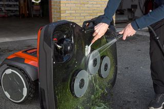 Eine Person, die die Unterseite eines Aufsitzmähers mit einem Schlauch reinigt, auf dem die Schneidmesser zu sehen sind.