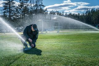 Ένα άτομο που ρυθμίζει έναν ψεκαστήρα σε ένα γήπεδο ποδοσφαίρου, που περιβάλλεται από ενεργούς ψεκαστήρες.