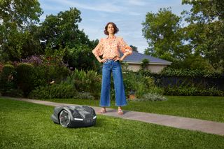 A person standing confidently in a garden with a robotic lawn mower on the grass.
