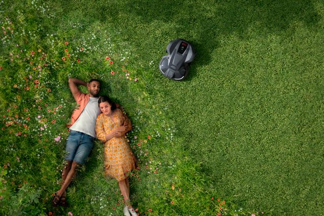 A couple lying on grass with a robotic lawn mower operating nearby.