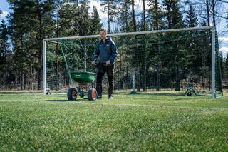 Ένα άτομο που χρησιμοποιεί έναν διανομέα γκαζόν σε ένα γήπεδο ποδοσφαίρου με ένα γκολπόστ στο παρασκήνιο.