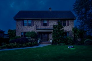 A robotic lawn mower operating on a lawn in front of a large brick house at night.