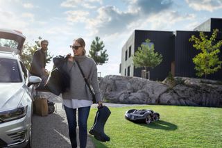 En person som bär på ett par stövlar och en väska, med en robotgräsklippare som arbetar på en gräsyta i närheten.