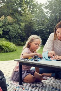 Una persona y un niño haciendo manualidades juntos en una mesa de patio con un robot cortacésped en el fondo.