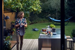 A person standing on a wooden deck next to an outdoor kitchen, with a robotic lawn mower operating on the lawn.