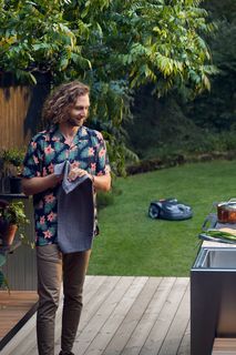 En person som står på ett trädäck bredvid ett utekök med en robotgräsklippare på gräsmattan.