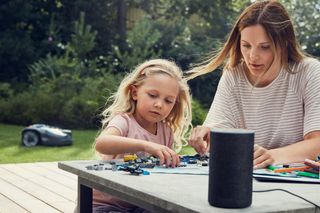 A child and an adult assembling toy blocks on a table outdoors, with a robotic lawn mower in the background.