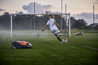 En person som spelar fotboll på en plan med två robotgräsklippare i närheten.
