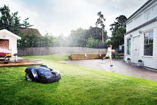 A robotic lawn mower operating in a garden during rain, with a person playing basketball nearby.