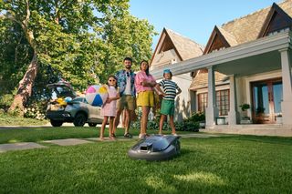 Un robot cortacésped funcionando en un césped frente a una casa, con una familia cerca.