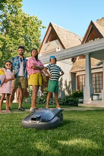 Ein Mähroboter arbeitet auf dem Rasen vor einem Haus, während eine Familie in der Nähe steht.