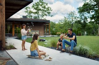 Família desfrutando de atividades ao ar livre em um jardim, com uma criança soprando bolhas e um robô corta-relva visível.