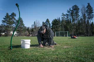 Ένα άτομο γονατιστό σε ένα χορτάρι, τείνει σε ένα κομμάτι γρασίδι με ένα κουβά και σωλήνα κοντά.