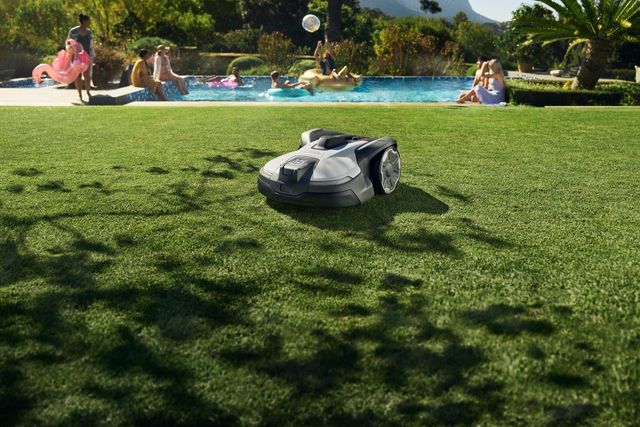 Robotic lawn mower operating on a well-maintained lawn near a swimming pool with people relaxing.