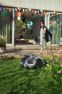 A robotic lawn mower operating in a garden with a party setup visible in the background.