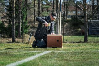A person kneeling on grass, using a drill on a wooden box in a fenced outdoor area.