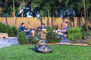 A robotic lawn mower operating on a lawn while three people sit around a fire pit in a garden.