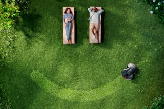 Deux personnes se détendent sur des chaises longues dans un jardin avec un robot tondeuse fonctionnant à proximité.