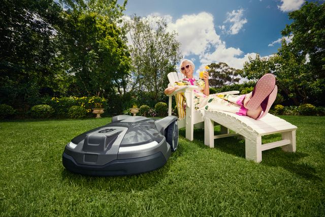 Ein Mähroboter in einem Garten, während eine Person es sich auf einer Sonnenliege entspannt.