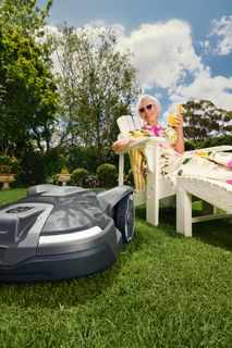 A robotic lawn mower operating in a garden while a person relaxes on a sun lounger.