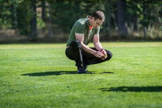 Uma pessoa ajoelhada em um campo gramado, examinando o chão de perto.