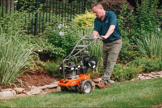A person using a garden tiller to cultivate soil along a landscaped garden edge.