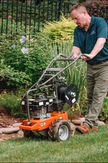 A person using a garden tiller to cultivate soil along a landscaped garden edge.