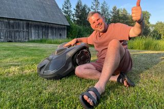 Cilvēks, kas sēž zālē blakus zāles pļāvēja robotam, dodot īkšķi uz augšu.