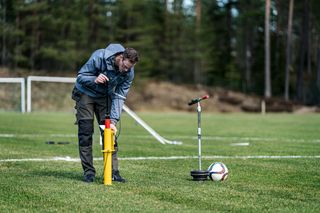 A person adjusting a yellow sports field line marker on a grassy field, with a soccer ball nearby.