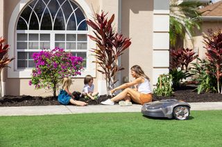 A robotic lawn mower operating near a family gardening in a residential yard.
