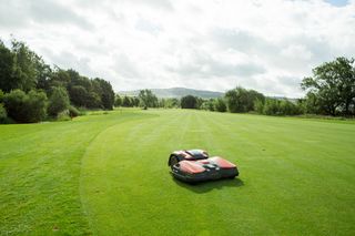 Robotic lawn mower operating on a large grassy field surrounded by trees.