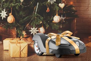 Ein mit einem goldenen Band umwickelter Husqvarna Mähroboter vor einem geschmückten Weihnachtsbaum.