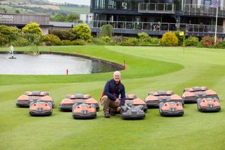 A person kneeling on a lawn surrounded by multiple robotic lawn mowers near a pond.
