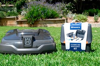 Husqvarna robotic lawn mower on grass with a cleaning and maintenance kit beside it.