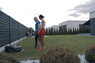 Du žmonės vaikšto modernioje sodo aplinkoje su robotu vejapjove, veikiančia ant žolės.