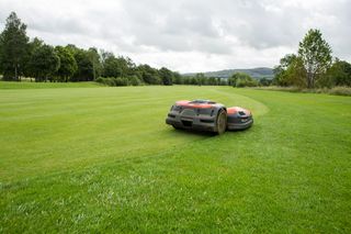 Robotic lawn mower operating on a large grassy field with trees in the background.