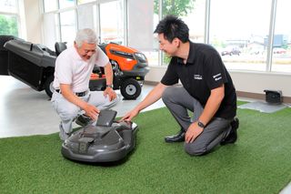 乗用芝刈り機を背景に、屋内でロボット芝刈り機を調べる2人。